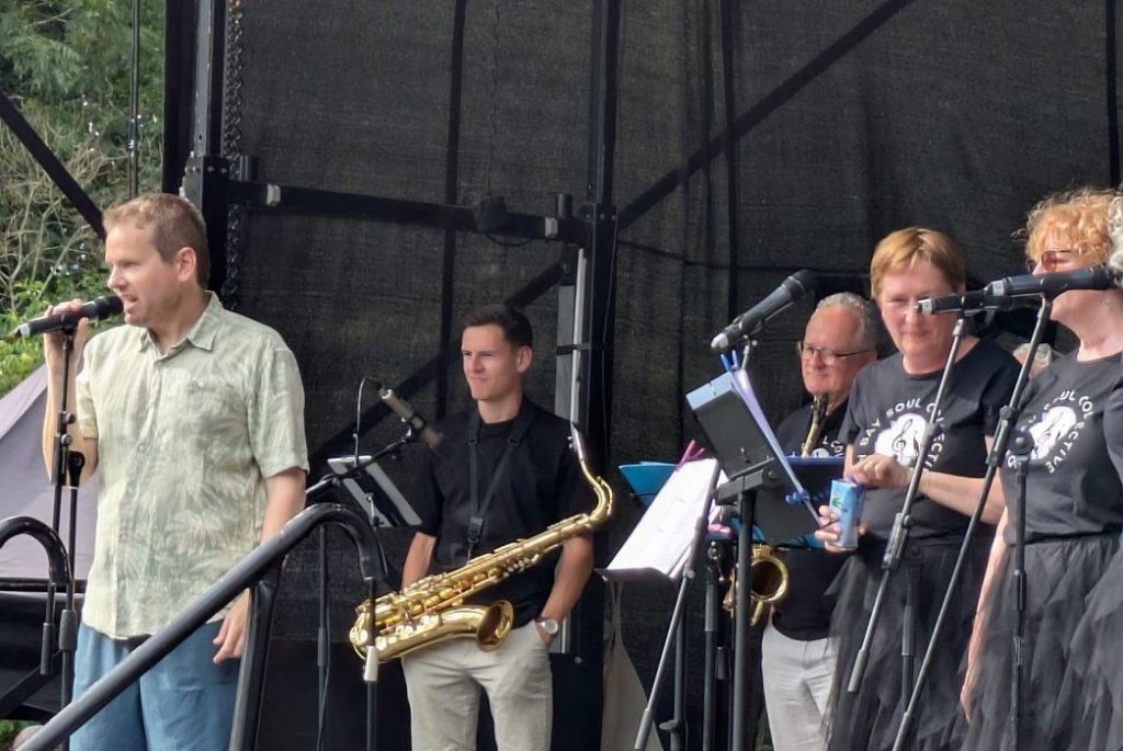 Conductor front of stage talking into microphone, singers and sax players in background